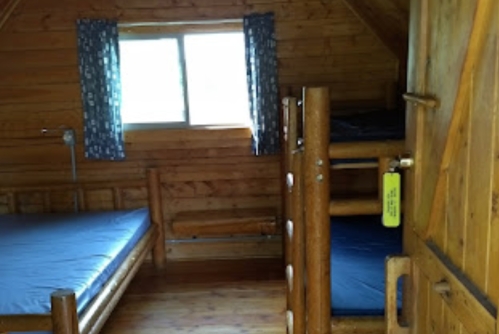 Cozy wooden cabin interior with two beds, a bunk bed, and a window with blue curtains.