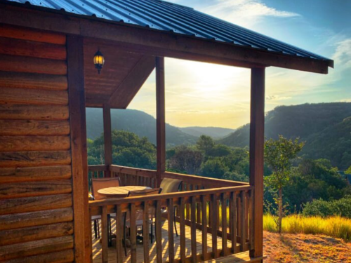 A wooden cabin porch overlooks a scenic valley at sunset, with mountains in the background and a clear sky.