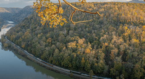 A scenic view of a river winding through a forested landscape with autumn foliage and a train along the bank.