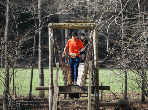 Two runners cross a wooden bridge in a forested area, surrounded by trees and a grassy field.