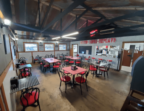 Interior of a casual restaurant featuring red and black tables and chairs, with a rustic decor and open kitchen area.