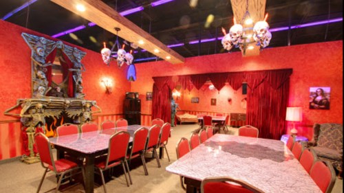 A themed room with red walls, skull decor, a long table, and chairs, featuring dramatic lighting and a cozy atmosphere.