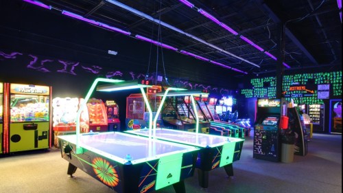 A vibrant arcade with neon lights, featuring air hockey tables and various colorful gaming machines.
