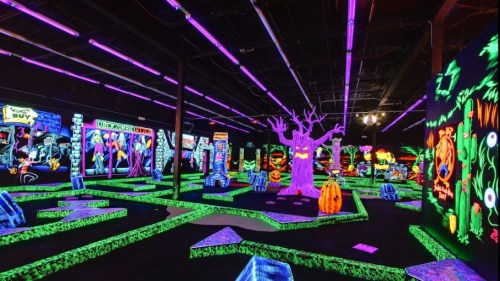 A vibrant glow-in-the-dark mini-golf course featuring neon decorations, spooky trees, and Halloween-themed elements.