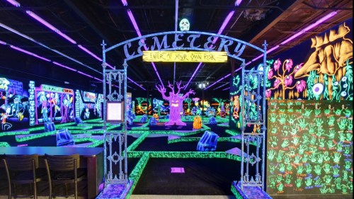 A vibrant glow-in-the-dark mini-golf course featuring a cemetery entrance, spooky decorations, and colorful neon elements.