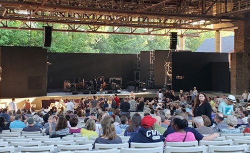 A crowd gathers in an outdoor amphitheater, with a stage set for a performance in the background.