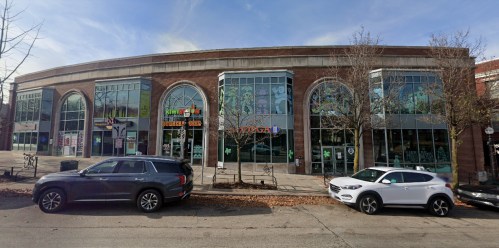 A large brick building with multiple arched windows, featuring shops and parked cars in front.