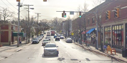 A busy street scene with cars, traffic lights, and shops lining the road on a cloudy day.