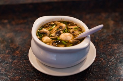 A white bowl of mushroom soup with green onions, resting on a small plate on a dark countertop.