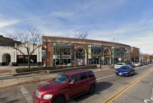 A street view of a commercial building with large windows, surrounded by trees and parked cars on a sunny day.