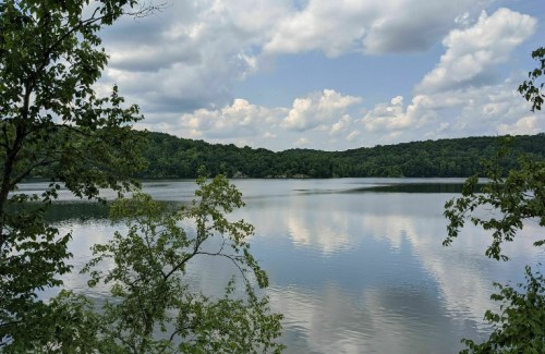 A serene lake surrounded by lush greenery and hills, reflecting fluffy clouds in the calm water.