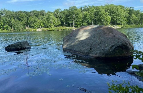 A large rock and a smaller boulder in a calm lake, surrounded by lush green trees under a clear blue sky.