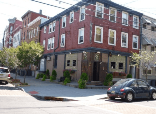 A red brick building with multiple windows, surrounded by greenery, on a sunny street with parked cars.