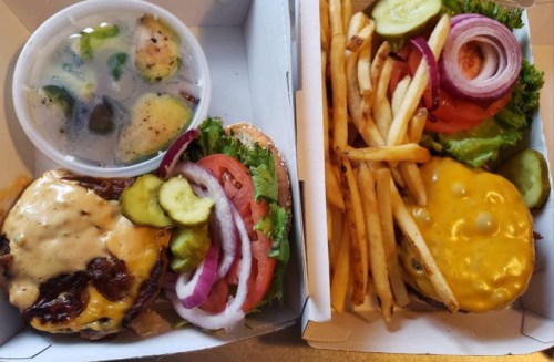 A burger with cheese, lettuce, tomato, pickles, and onions, served with fries and a cup of soup on the side.