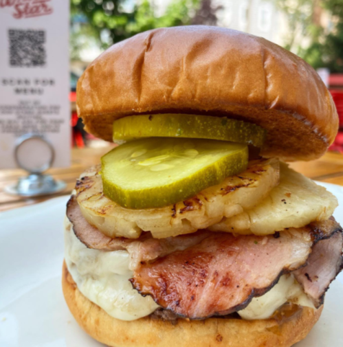 A close-up of a sandwich with ham, melted cheese, grilled pineapple, and pickles on a toasted bun.