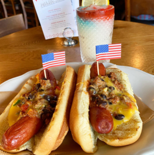 Two hot dogs in buns topped with chili, cheese, and small American flags, served with a colorful drink in the background.