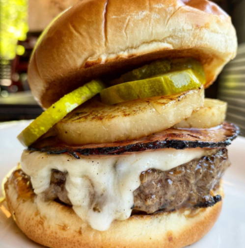 A close-up of a burger with a juicy patty, melted cheese, grilled pineapple, and pickles on a toasted bun.