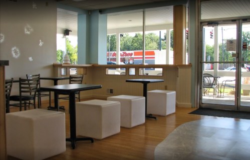 A bright, modern café interior with tables, chairs, and white cube seating near large windows.