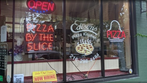 A storefront window featuring neon signs for "Edison's Pizza Kitchen," advertising pizza by the slice and hiring.