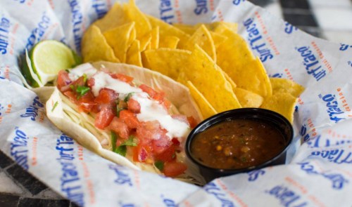 A plate featuring a taco with fresh toppings, tortilla chips, a lime wedge, and a small bowl of salsa.