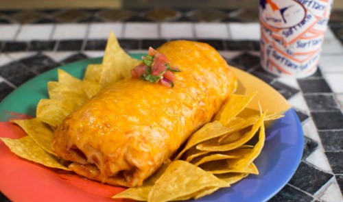 A large, cheesy burrito topped with salsa, served on a colorful plate with tortilla chips and a drink in the background.