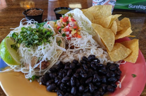 A colorful plate featuring tacos with toppings, rice, black beans, tortilla chips, and lime wedges.