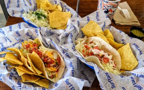 Two plates of tacos with toppings, tortilla chips, and a side of salsa on a table lined with branded paper.
