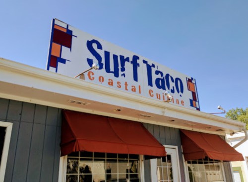 Sign for "Surf Taco" with the tagline "Coastal Cuisine" above a restaurant entrance with red awnings.