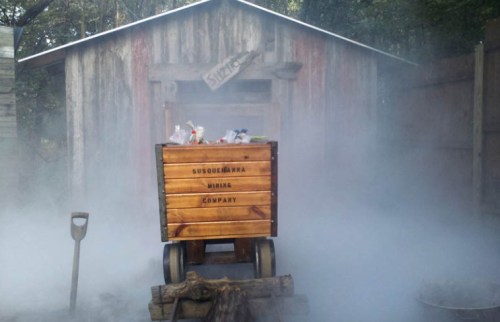 A wooden cart labeled "Susquehanna Mining Company" sits in fog near a rustic shed in a wooded area.