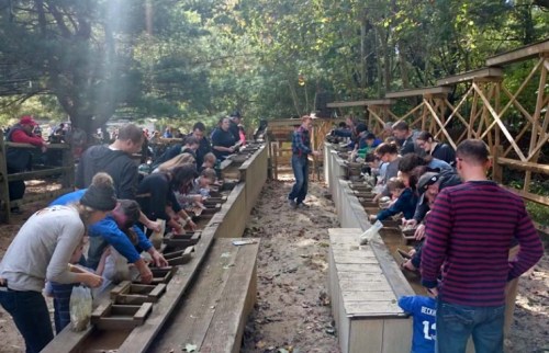 A crowd of people sifting through sand at outdoor tables, searching for gems or fossils in a wooded area.