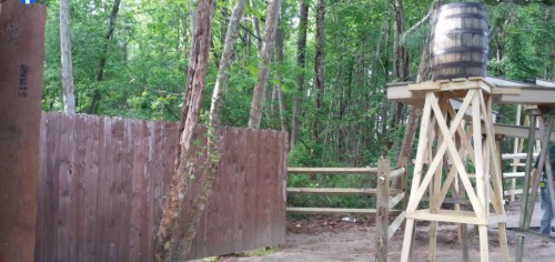 A wooden fence and a raised platform with a barrel, surrounded by trees in a forested area.