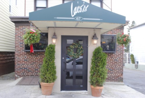 Entrance of a restaurant with a green awning, festive wreath, and potted plants on either side.