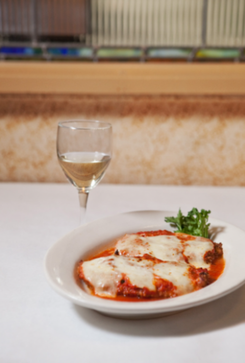 A plate of lasagna with marinara sauce and melted cheese, accompanied by a glass of white wine and a sprig of parsley.