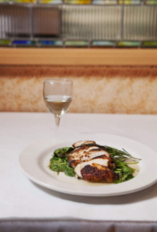A plate of grilled chicken on a bed of greens, accompanied by a glass of white wine, set on a white tablecloth.