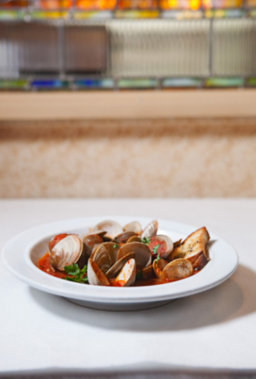 A white bowl filled with clams in a rich tomato sauce, garnished with herbs, on a white tablecloth.