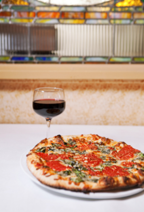 A freshly baked pizza topped with sauce and greens, accompanied by a glass of red wine, set against a stained glass backdrop.