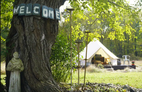 A tree with a "WELCOME" sign, a statue, and a glamping tent in a serene outdoor setting.