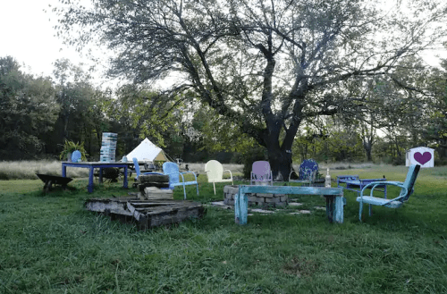 A rustic outdoor seating area with colorful chairs and tables under a large tree, surrounded by greenery.