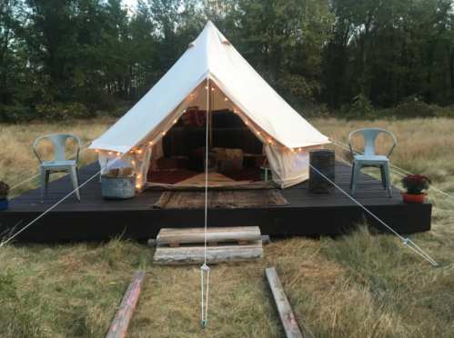 A cozy glamping tent on a wooden platform, surrounded by grass, with string lights and chairs outside.