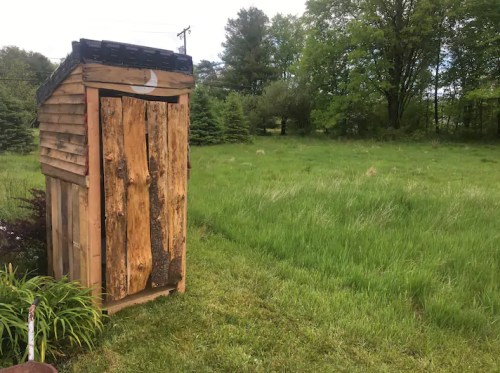 A rustic wooden outhouse stands in a grassy field surrounded by trees under a cloudy sky.