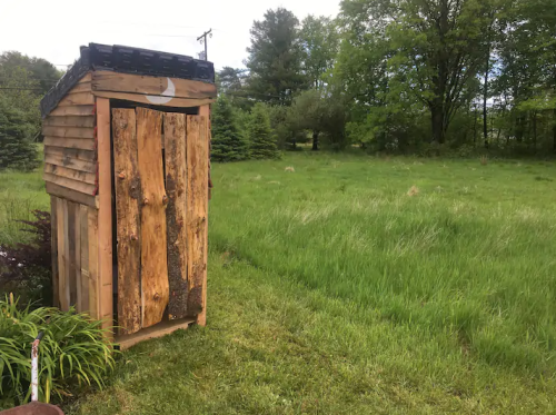 A rustic wooden outhouse stands in a grassy field surrounded by trees under a cloudy sky.