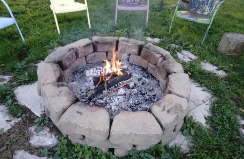 A stone fire pit with a small fire burning, surrounded by grassy area and colorful chairs in the background.