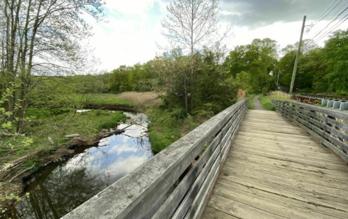 A wooden bridge over a calm stream, surrounded by lush greenery and trees under a partly cloudy sky.