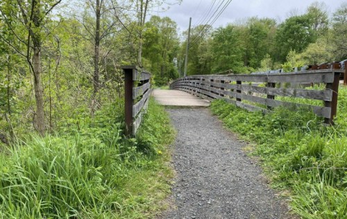 A gravel path leads to a wooden bridge surrounded by lush greenery and trees under a cloudy sky.