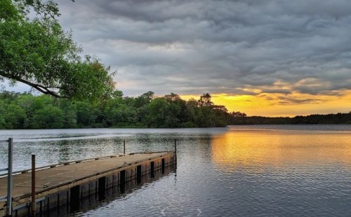 A serene lake at sunset, with a wooden dock extending into the water and clouds reflecting warm colors in the sky.
