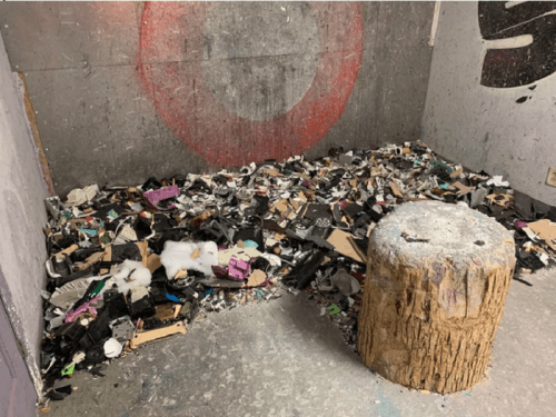A cluttered room floor covered with various debris, including plastic, cardboard, and foam, next to a wooden stump.