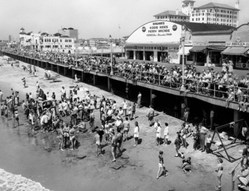 A crowded beach scene with people enjoying the water and a bustling boardwalk in the background, captured in black and white.