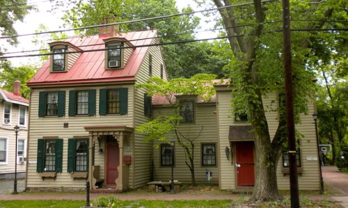 A two-story house with a red metal roof, green shutters, and a tree in the front yard, surrounded by greenery.
