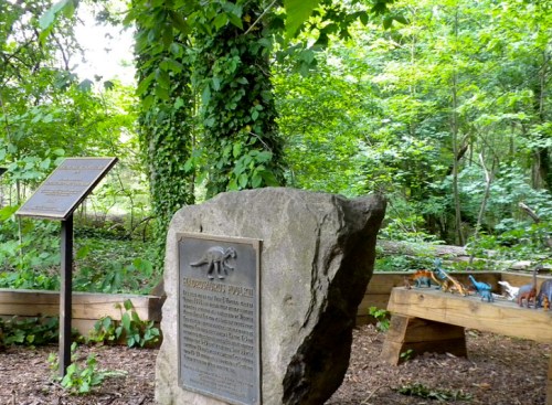 A stone monument in a wooded area, with informational plaques and small dinosaur figures on a wooden display.