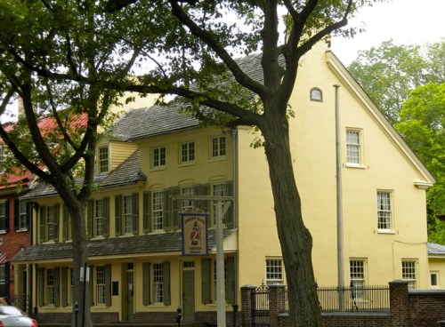 Historic yellow building with multiple windows, surrounded by trees, featuring a sign hanging from a post.